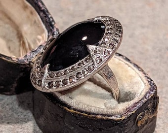 May include: An oval black gemstone ring with a silver-colored setting, displayed in a vintage black jewelry box. The ring's setting is adorned with small, sparkling accents. The box has a cream-colored interior.