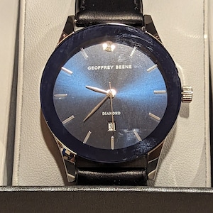 May include: A silver and black watch with a blue face and a black leather band. The watch face has a diamond at the 12 o'clock position and the text "GEOFFREY BEENE" and "DIAMOND" printed on it.