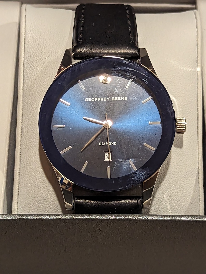 May include: A silver and black watch with a blue face and a black leather band. The watch face has a diamond at the 12 o'clock position and the text "GEOFFREY BEENE" and "DIAMOND" printed on it.