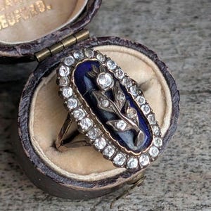 May include: An antique gold ring with an oval, dark blue enamel center featuring a floral design. The ring is surrounded by a border of small, clear gemstones and is displayed in a vintage jewelry box.