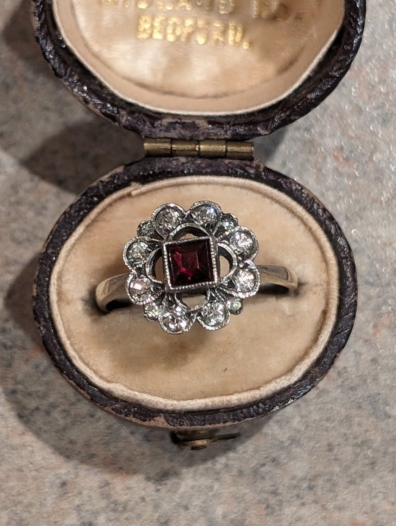 Pu&ograve; includere: Un anello color argento con una gemma rosso scuro a taglio quadrato al centro, circondata da un alone floreale di piccole pietre rotonde trasparenti. L'anello &egrave; esposto all'interno di un portagioie ovale marrone scuro con interno color crema.
