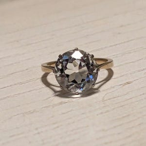 May include: A gold ring with a large, clear, oval-shaped gemstone. The gemstone has many facets and is set in a simple, gold band.