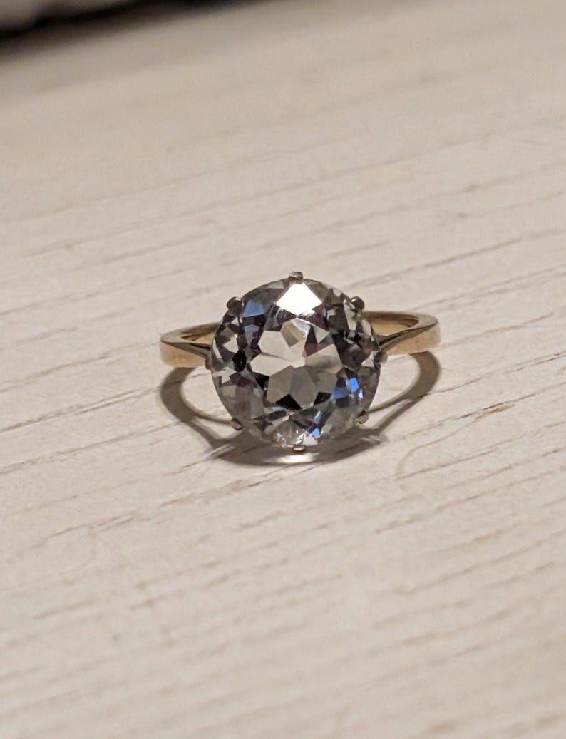 May include: A gold ring with a large, clear, oval-shaped gemstone. The gemstone has many facets and is set in a simple, gold band.