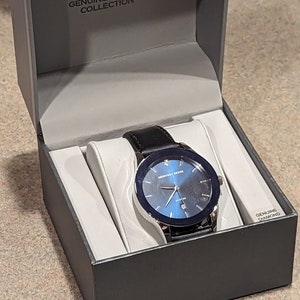 May include: A black leather watch with a blue face and silver trim. The watch is in a black box with the words "Geoffrey Beene Genuine Diamond Collection" on the top. The box also has the words "Genuine Diamond" on the side.