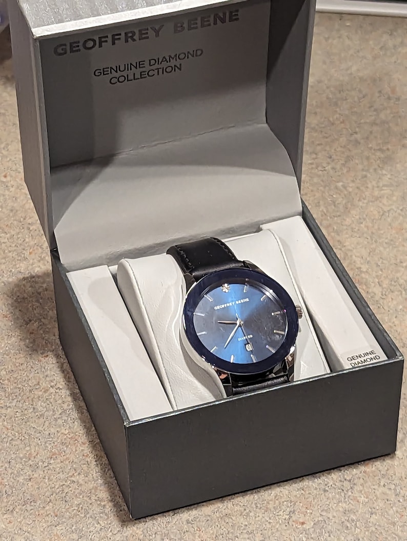 May include: A black leather watch with a blue face and silver trim. The watch is in a black box with the words "Geoffrey Beene Genuine Diamond Collection" on the top. The box also has the words "Genuine Diamond" on the side.