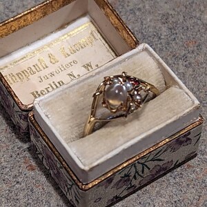 K&ouml;nnte beinhalten: Ein goldener Ring mit einem gro&szlig;en, runden, perlenartigen Mittelstein und kleinen roten Akzenten, pr&auml;sentiert in einer offenen, floralen Vintage-Schmuckschatulle. Die Schatulle hat einen goldenen Rand und den Text "H&uuml;ppauf & Kumpf Juwoliere Berlin N. W."