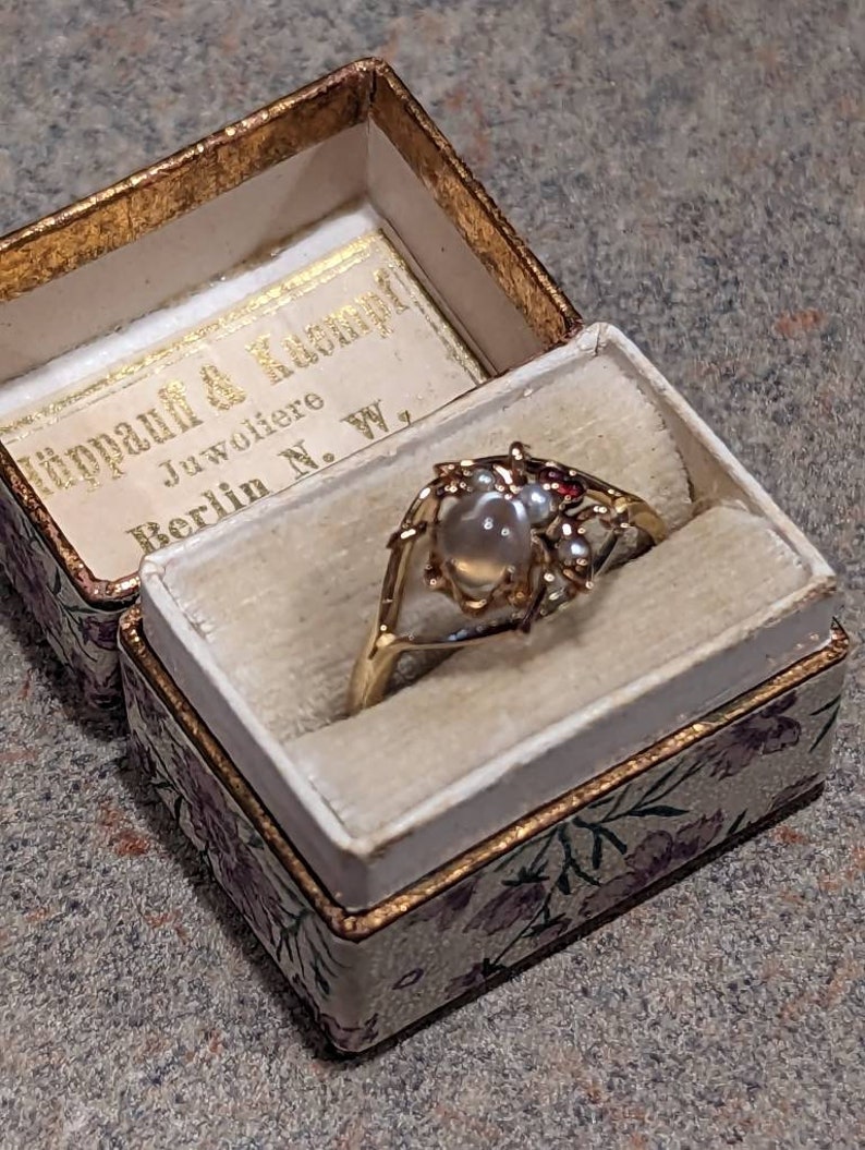 K&ouml;nnte beinhalten: Ein goldener Ring mit einem gro&szlig;en, runden, perlenartigen Mittelstein und kleinen roten Akzenten, pr&auml;sentiert in einer offenen, floralen Vintage-Schmuckschatulle. Die Schatulle hat einen goldenen Rand und den Text "H&uuml;ppauf & Kumpf Juwoliere Berlin N. W."