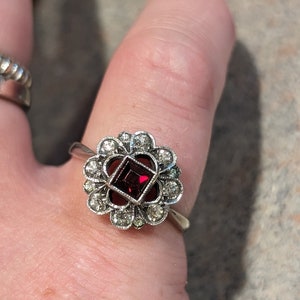 Pu&ograve; includere: Un anello color argento con una pietra preziosa centrale rosso scuro a taglio quadrato, circondata da un'aureola a forma di fiore composta da piccole pietre chiare a taglio rotondo. La montatura ha un bordo testurizzato e il gambo &egrave; liscio.