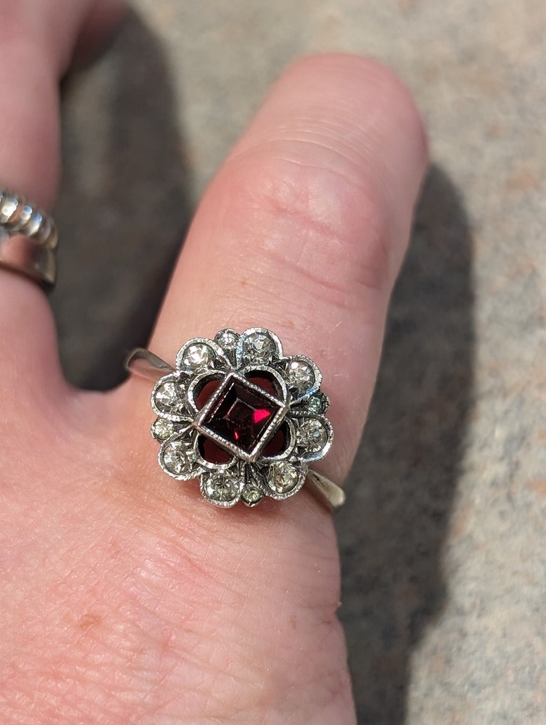 Pu&ograve; includere: Un anello color argento con una pietra preziosa centrale rosso scuro a taglio quadrato, circondata da un'aureola a forma di fiore composta da piccole pietre chiare a taglio rotondo. La montatura ha un bordo testurizzato e il gambo &egrave; liscio.