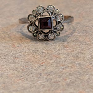 Pu&ograve; includere: Un anello color argento con una pietra preziosa rosso scuro a taglio quadrato al centro, circondata da un'aureola a forma di fiore composta da piccole pietre chiare a taglio rotondo incastonate in una montatura decorativa.