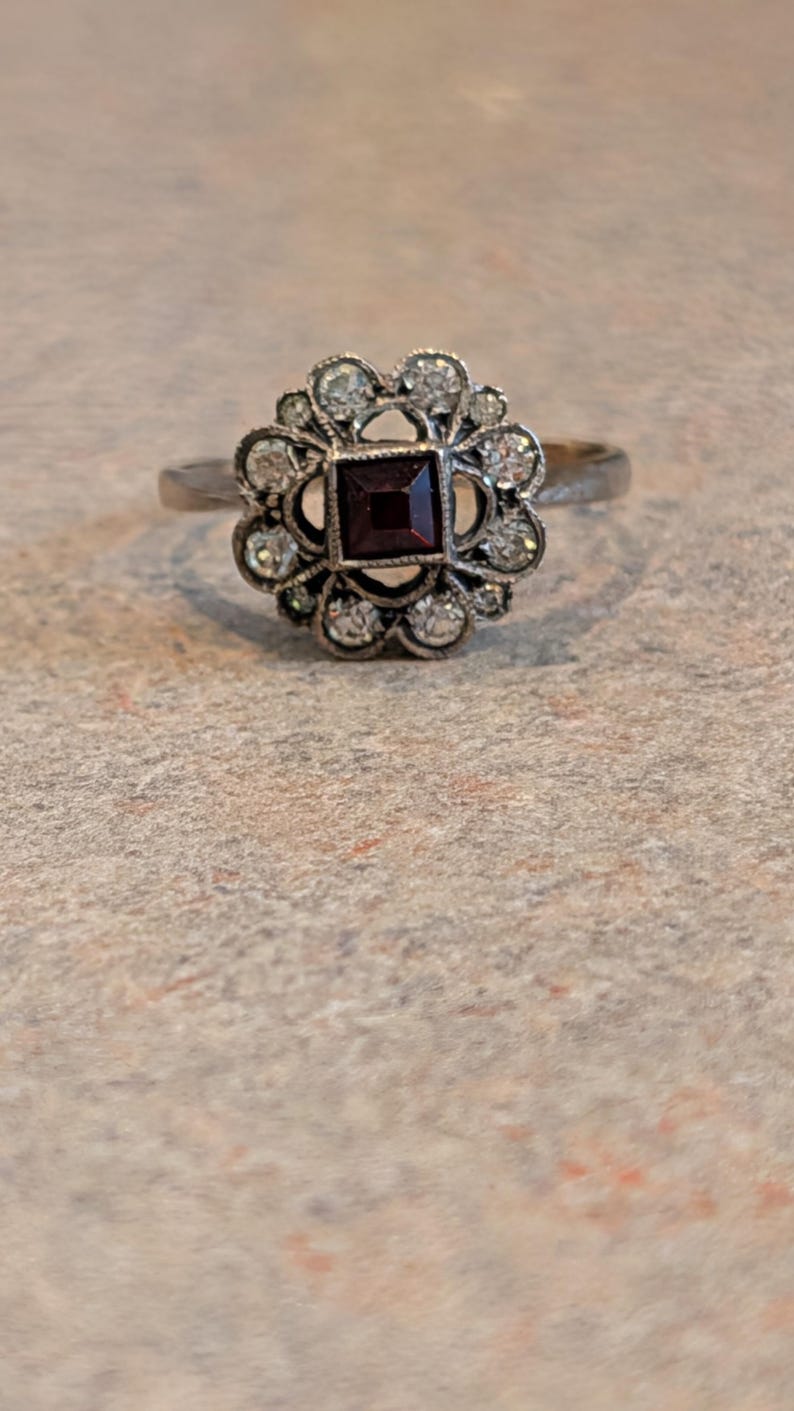 Pu&ograve; includere: Un anello color argento con una pietra preziosa rosso scuro a taglio quadrato al centro, circondata da un'aureola a forma di fiore composta da piccole pietre chiare a taglio rotondo incastonate in una montatura decorativa.