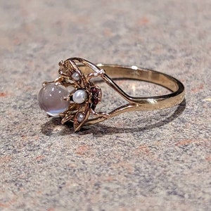 K&ouml;nnte beinhalten: Ein goldener Ring mit einem gro&szlig;en, runden, grauen Edelstein, kleinen Perlen und roten Edelsteinen. Der Ring hat ein kompliziertes, blumeninspiriertes Design um den Hauptstein mit einem zarten Band.