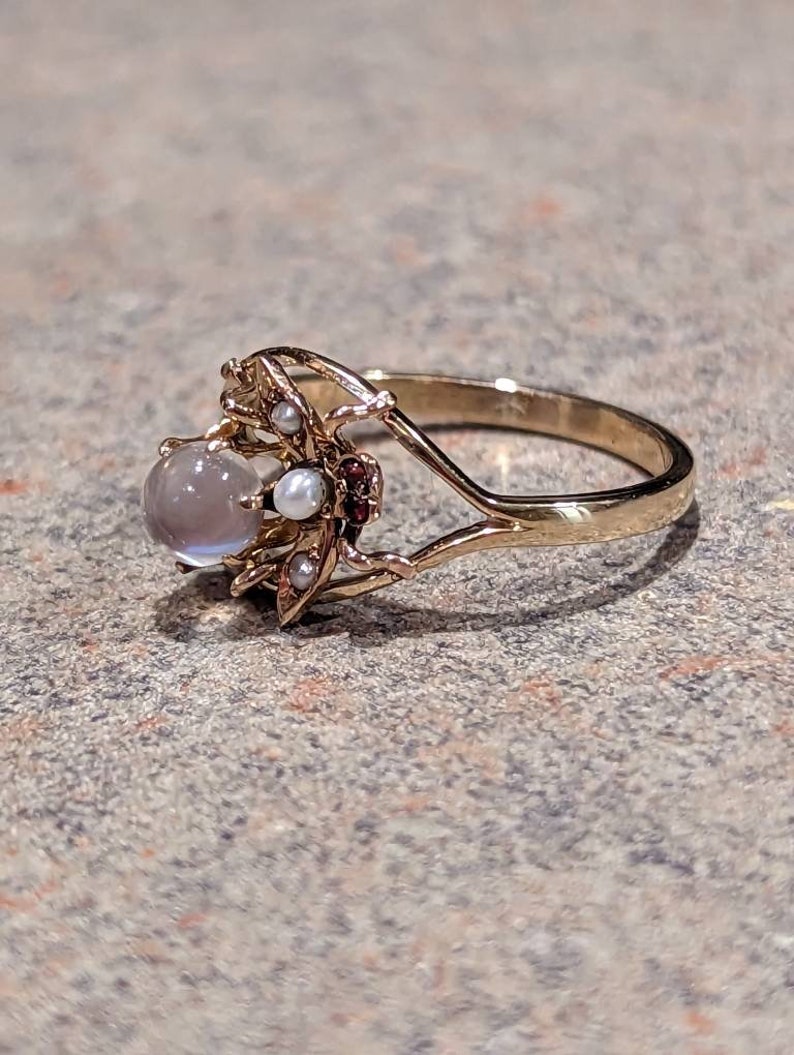 K&ouml;nnte beinhalten: Ein goldener Ring mit einem gro&szlig;en, runden, grauen Edelstein, kleinen Perlen und roten Edelsteinen. Der Ring hat ein kompliziertes, blumeninspiriertes Design um den Hauptstein mit einem zarten Band.