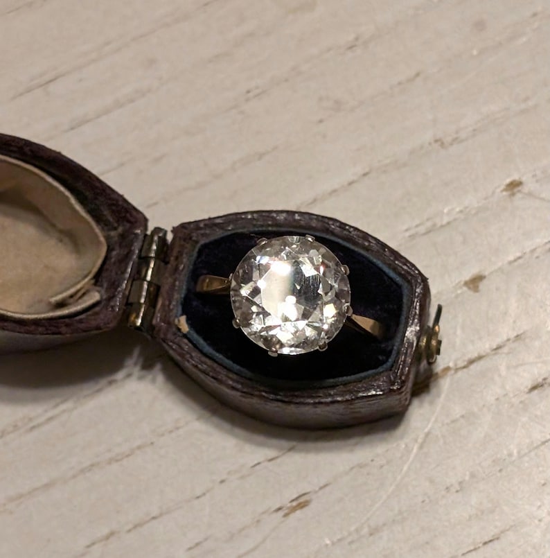 May include: A clear, round gemstone ring set in a gold band. The ring is in a small, brown velvet ring box.