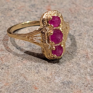 May include: A gold-colored ring featuring three oval-cut, deep red gemstones set vertically. The ring has an ornate, textured design around the gemstones and on the band. The ring is displayed on a textured, gray surface.