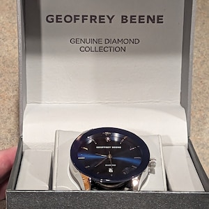 May include: A silver watch with a blue face and a diamond in the center. The watch is in a box that says "Geoffrey Beene Genuine Diamond Collection".