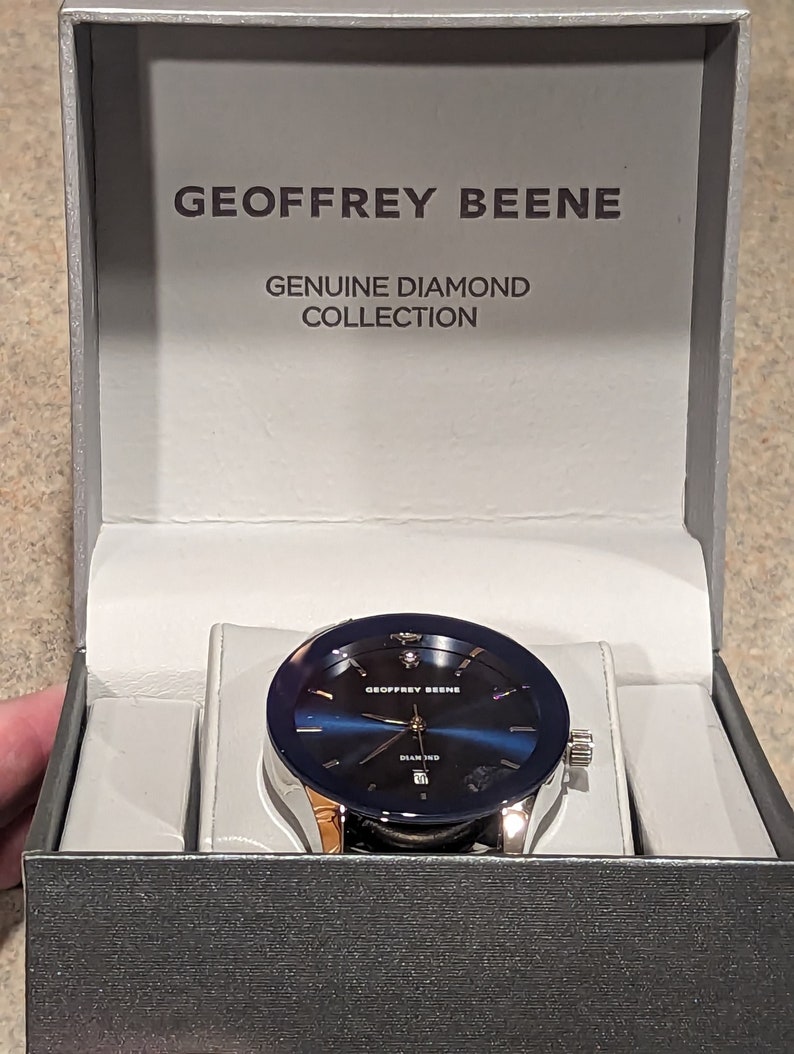 May include: A silver watch with a blue face and a diamond in the center. The watch is in a box that says "Geoffrey Beene Genuine Diamond Collection".