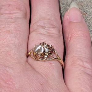 K&ouml;nnte beinhalten: Ein goldfarbener Ring mit einem gro&szlig;en, ovalen, milchig-wei&szlig;en Stein in der Mitte. Der Ring hat ein verziertes Design mit kleineren Perlen und roten Akzenten. Der Ring befindet sich an einem Finger.