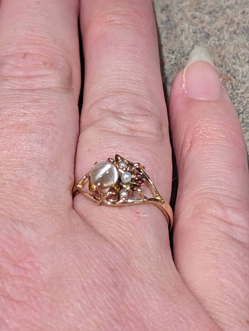 K&ouml;nnte beinhalten: Ein goldfarbener Ring mit einem gro&szlig;en, ovalen, milchig-wei&szlig;en Stein in der Mitte. Der Ring hat ein verziertes Design mit kleineren Perlen und roten Akzenten. Der Ring befindet sich an einem Finger.