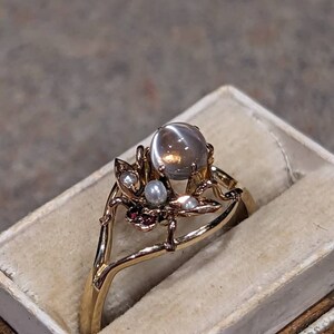 K&ouml;nnte beinhalten: Ein goldener Ring mit einem gro&szlig;en, runden, grauen Edelstein als Mittelpunkt. Der Ring weist filigrane Blumen- und Zweig-Designs mit kleinen Perlen und roten Akzenten auf. Der Ring wird in einer cremefarbenen Schmuckschatulle pr&auml;sentiert.