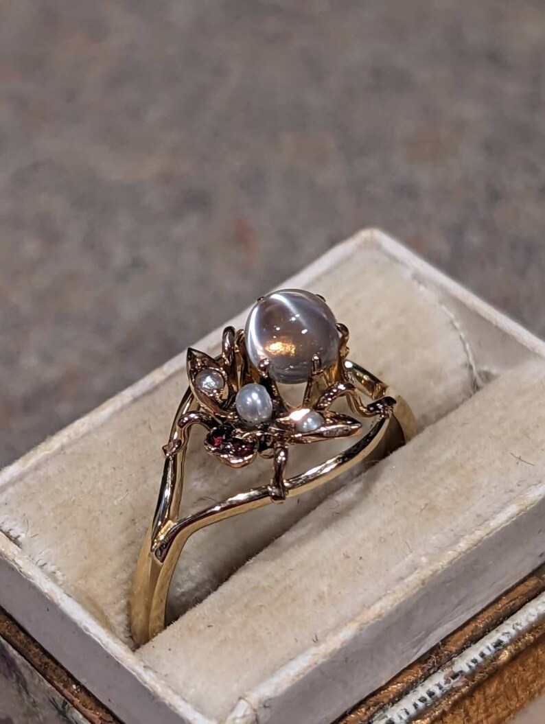 K&ouml;nnte beinhalten: Ein goldener Ring mit einem gro&szlig;en, runden, grauen Edelstein als Mittelpunkt. Der Ring weist filigrane Blumen- und Zweig-Designs mit kleinen Perlen und roten Akzenten auf. Der Ring wird in einer cremefarbenen Schmuckschatulle pr&auml;sentiert.
