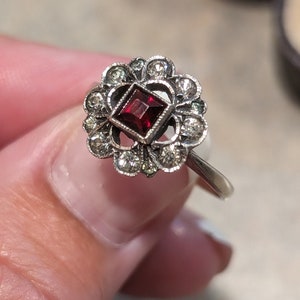 Pu&ograve; includere: Un anello color argento con una pietra preziosa centrale rosso scuro a taglio quadrato. La pietra &egrave; circondata da un'aureola a forma di fiore composta da piccole pietre rotonde e trasparenti incastonate in una montatura decorativa.