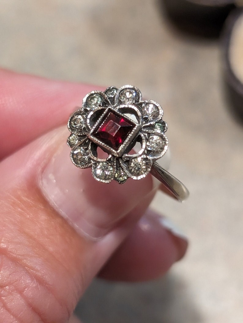 Pu&ograve; includere: Un anello color argento con una pietra preziosa centrale rosso scuro a taglio quadrato. La pietra &egrave; circondata da un'aureola a forma di fiore composta da piccole pietre rotonde e trasparenti incastonate in una montatura decorativa.