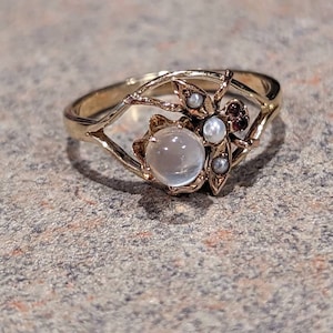 K&ouml;nnte beinhalten: Ein goldfarbener Ring mit einem zentralen, runden, durchscheinenden Stein. Der Ring hat ein insektenartiges Design mit kleinen Perlen und roten Edelsteinen. Das Band ist d&uuml;nn und filigran, mit einem durchbrochenen Design um den zentralen Stein.