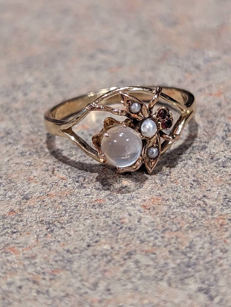 K&ouml;nnte beinhalten: Ein goldfarbener Ring mit einem zentralen, runden, durchscheinenden Stein. Der Ring hat ein insektenartiges Design mit kleinen Perlen und roten Edelsteinen. Das Band ist d&uuml;nn und filigran, mit einem durchbrochenen Design um den zentralen Stein.