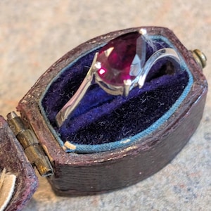 May include: A silver ring with a pear-shaped, deep red gemstone, presented in an open, antique, purple jewellery box with a blue velvet interior. The ring's band curves elegantly around the gemstone.