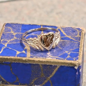 以下が含まれることがあります： ハート型のセッティングに金の「M」の文字が施された銀の指輪。指輪は青と金の模様の箱の上にある。