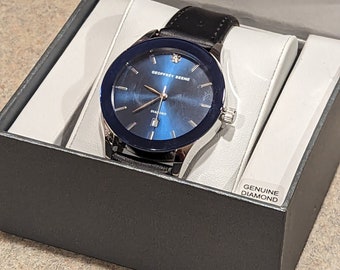 Könnte beinhalten: Eine Armbanduhr aus schwarzem Leder mit einem silbernen Gehäuse und einem blauen Zifferblatt. Die Uhr hat einen Diamanten auf dem Zifferblatt und die Worte "Geoffrey Beene" und "Diamond" sind auf dem Zifferblatt aufgedruckt. Die Uhr befindet sich in einer schwarzen Box mit der Aufschrift "Genuine Diamond" auf der Box.