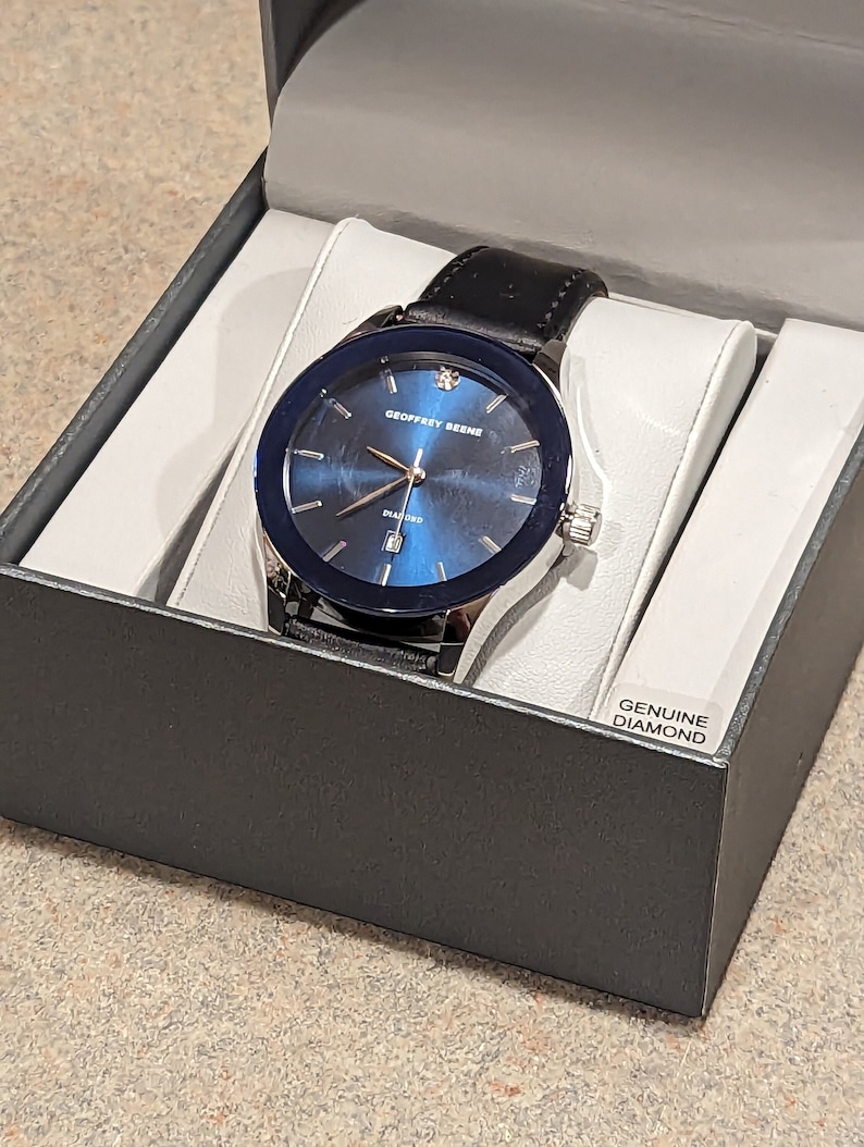 May include: A black leather wristwatch with a silver case and a blue face. The watch has a diamond on the face and the words "Geoffrey Beene" and "Diamond" printed on the face. The watch is in a black box with the words "Genuine Diamond" printed on the box.