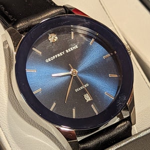 May include: A silver and black watch with a blue face and a black leather band. The watch face has a diamond at the 12 o'clock position and the words "GEOFFREY BEENE" and "DIAMOND" printed on it.