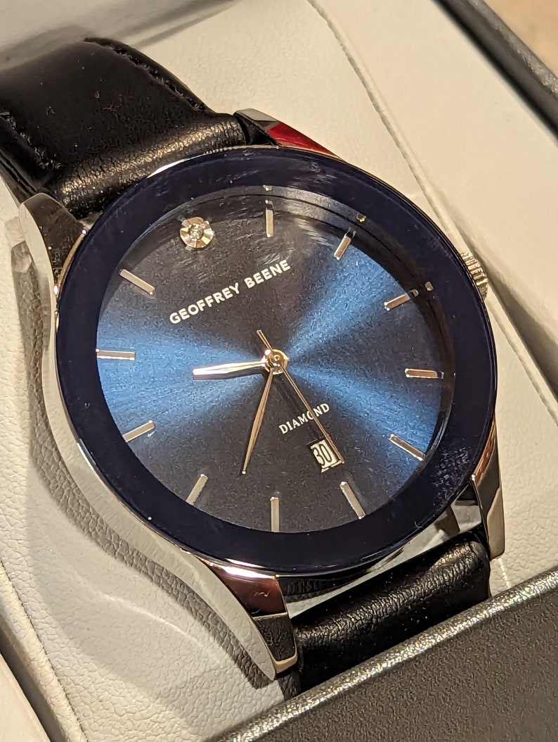 May include: A silver and black watch with a blue face and a black leather band. The watch face has a diamond at the 12 o'clock position and the words "GEOFFREY BEENE" and "DIAMOND" printed on it.