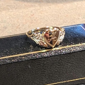 May include: A gold ring with a heart-shaped design featuring a letter 'L' inside. The ring is set in a black box.