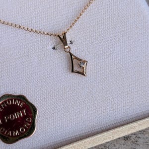 May include: A gold-toned necklace with a diamond pendant in a diamond shape. The pendant is centered on a delicate chain. The necklace is presented in a cream-colored jewelry box, with a gold-colored seal visible.