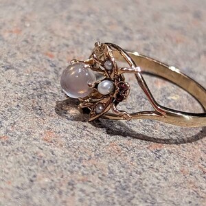 K&ouml;nnte beinhalten: Ein goldfarbener Ring mit einem runden, hellen Edelstein und zarten floralen und Perlenakzenten. Der Ring hat ein schmales Band und die Fassung kleine Perlen und rotbraune Steine.