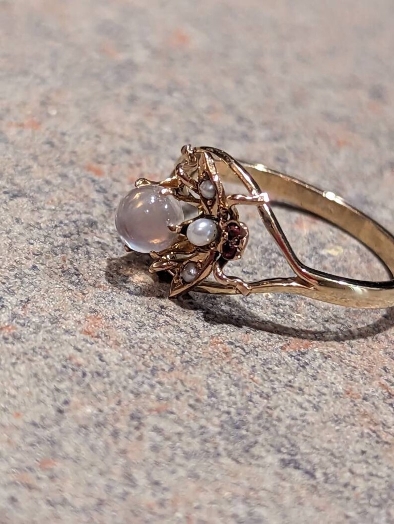 K&ouml;nnte beinhalten: Ein goldfarbener Ring mit einem runden, hellen Edelstein und zarten floralen und Perlenakzenten. Der Ring hat ein schmales Band und die Fassung kleine Perlen und rotbraune Steine.