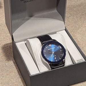 May include: A black leather band watch with a silver case and a blue face. The watch has a single diamond on the bezel and the words "Geoffrey Beene" and "Genuine Diamond Collection" on the face. The watch is in a black box with a white interior.