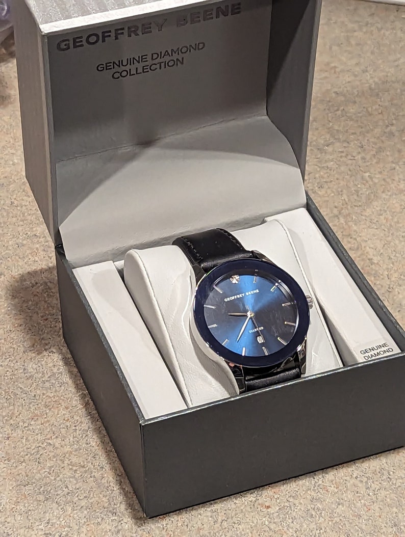May include: A black leather band watch with a silver case and a blue face. The watch has a single diamond on the bezel and the words "Geoffrey Beene" and "Genuine Diamond Collection" on the face. The watch is in a black box with a white interior.