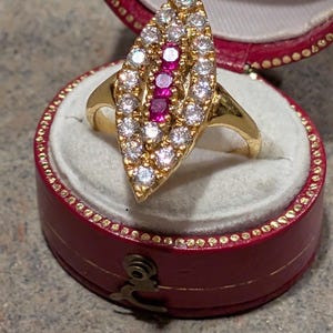 Könnte beinhalten: Ein goldener Ring mit einer markisenförmigen Fassung, besetzt mit weißen und roten Edelsteinen. Der Ring wird in einer roten Schmuckschatulle mit weißem Innenraum präsentiert.