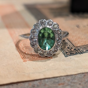 May include: A silver ring with a central oval-cut green gemstone, surrounded by a halo of smaller clear stones. The ring is set on a vintage paper background, showcasing the jewelry's design and color.