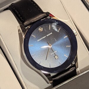 May include: A silver watch with a black leather band and a blue face. The watch has a diamond on the face and the words "Geoffrey Beene Diamond" printed on the face.
