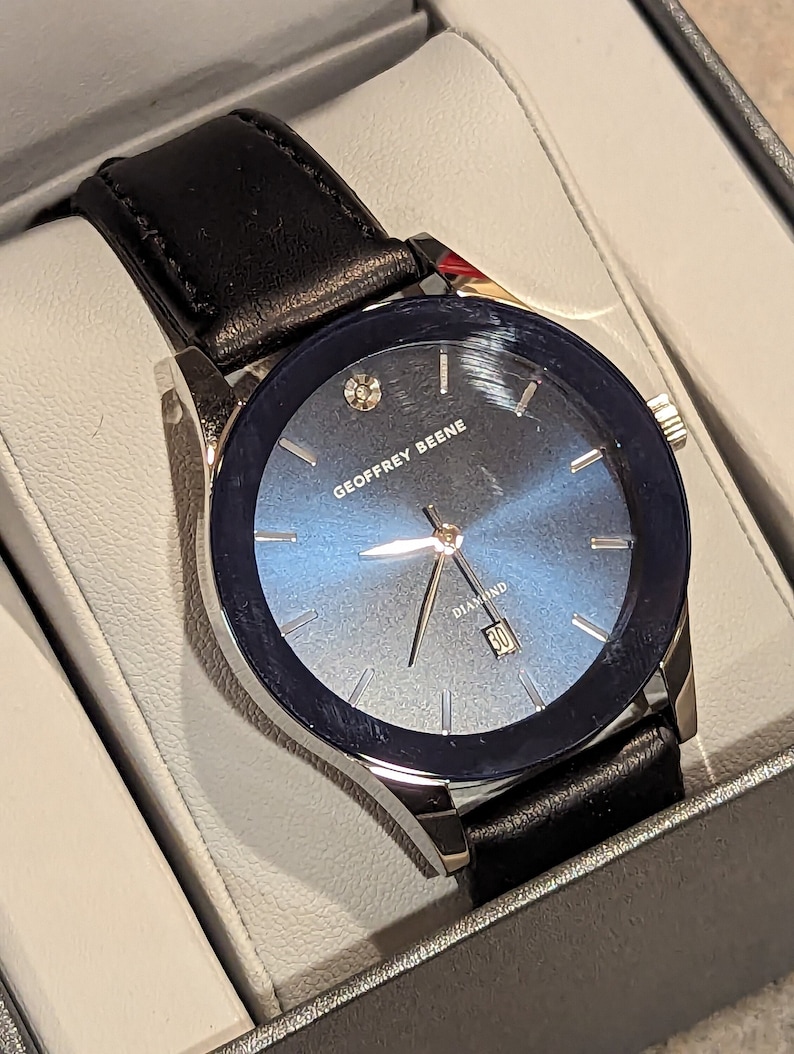 May include: A silver watch with a black leather band and a blue face. The watch has a diamond on the face and the words "Geoffrey Beene Diamond" printed on the face.