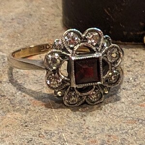 Pu&ograve; includere: Un anello in oro con una pietra preziosa rosso scuro a taglio quadrato al centro, circondata da un gruppo di piccole pietre rotonde e chiare a forma di fiore, incastonate in una montatura in metallo argentato. Il gambo &egrave; sottile e dorato.