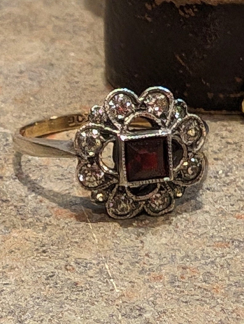 Pu&ograve; includere: Un anello in oro con una pietra preziosa rosso scuro a taglio quadrato al centro, circondata da un gruppo di piccole pietre rotonde e chiare a forma di fiore, incastonate in una montatura in metallo argentato. Il gambo &egrave; sottile e dorato.