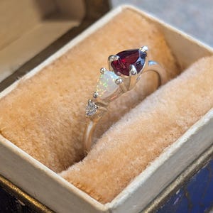May include: A silver ring with a pear-shaped, deep red gemstone and a white opal stone. The ring is set with small accent stones and is displayed in a vintage blue and gold jewelry box with a peach-colored interior.