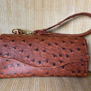May include: A brown wallet with a wrist strap. The wallet has a textured surface with dark brown spots. It features a curved flap closure and a gold-colored clasp. The wrist strap is made of the same material as the wallet.