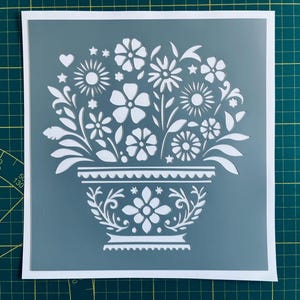 May include: A white stencil of a flower pot with a floral design. The pot has a scalloped rim and a decorative pattern. The flowers are stylised and have a simple design.
